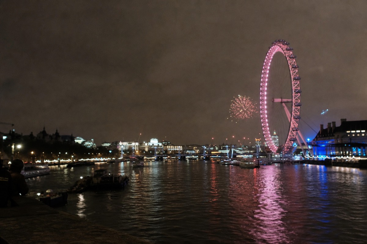 A night skyline over the Thames with firework glow reflected on the water near the illuminated London Eye