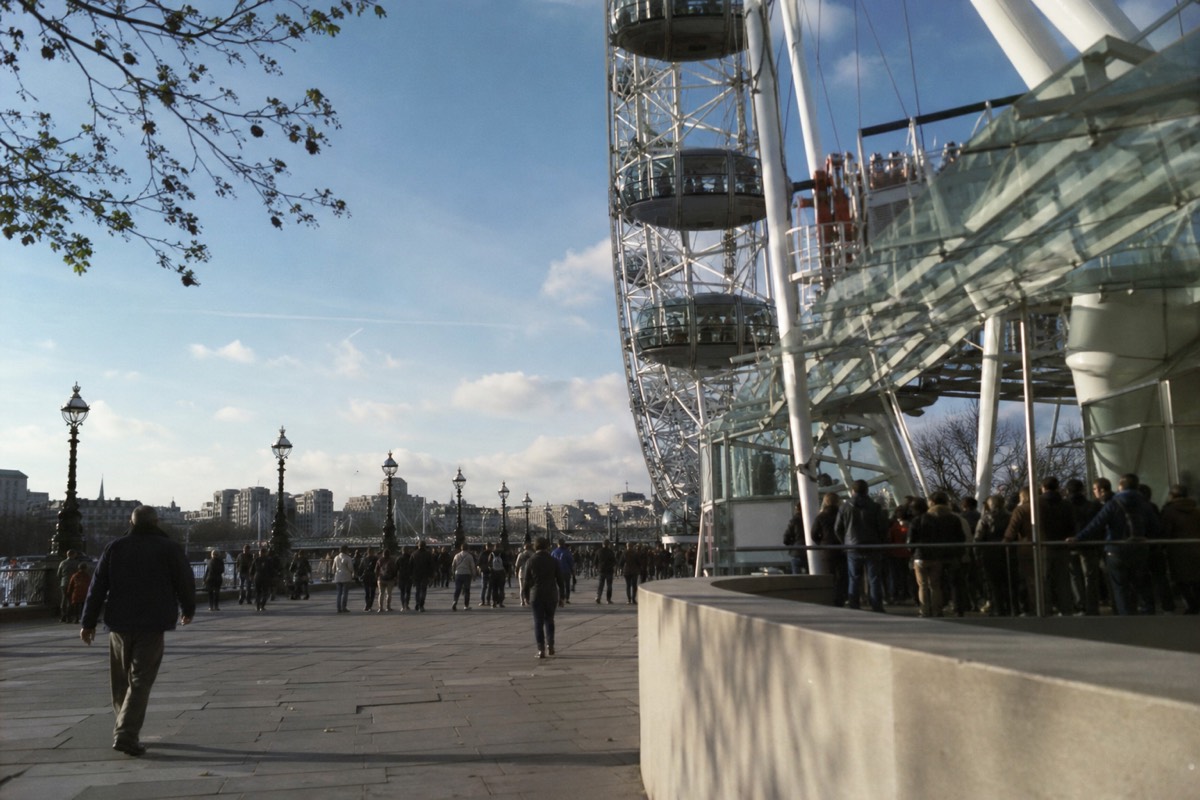 A crowded riverside plaza with visitors queueing near the base of the London Eye
